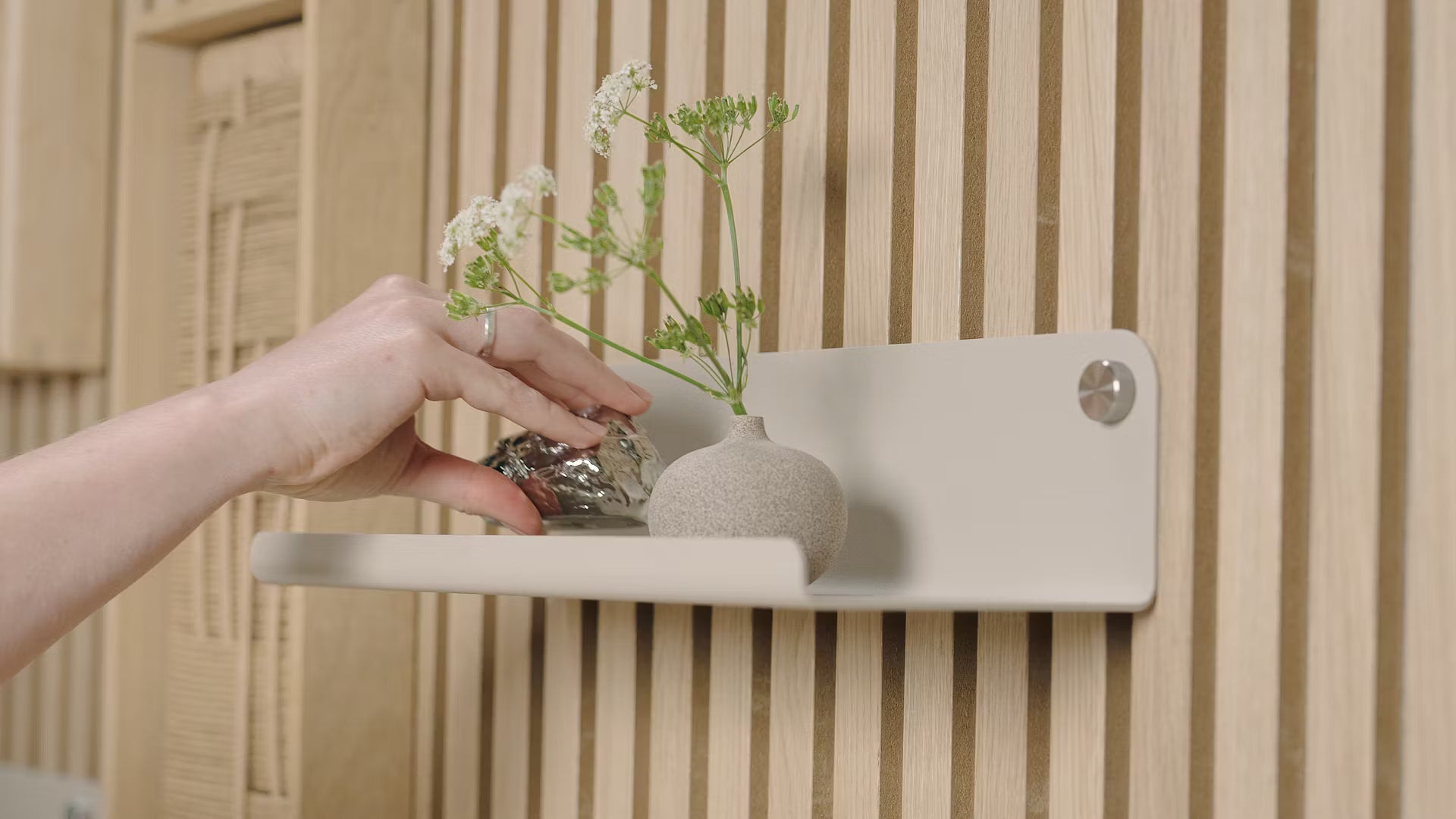 Eine Hand platziert ein kleines dekoratives Objekt neben einer Vase mit weißen Blumen auf einem minimalistischen Regal, das vor einer strukturierten nordischen Akustikholz-Lamellenwand steht.